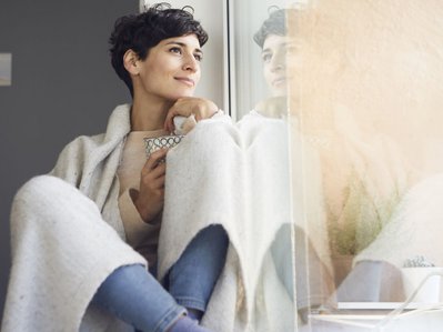 Eine in eine Decke eingekuschelte Frau sitzt am Fenster mit ihrer Tasse.  Eine Heizungsinstallation sorgt für effiziente, zuverlässige Wärme.