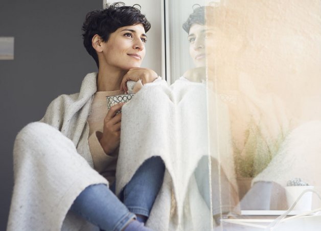 Eine in eine Decke eingekuschelte Frau sitzt am Fenster mit ihrer Tasse.  Eine Heizungsinstallation sorgt für effiziente, zuverlässige Wärme.