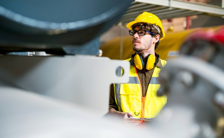 Ingenieur mit Schutzhelm und reflektierender Weste arbeitet in einer Fabrikhalle. Konzentration und Sicherheit stehen im Vordergrund.