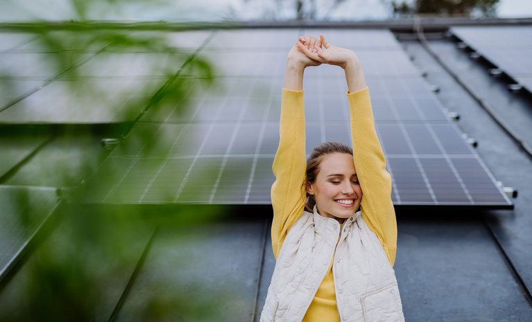 Frau in gelbem Pullover streckt sich lächelnd vor einer Solarzellenwand. Blätter im Vordergrund, Symbol für nachhaltige Energie und positive Ausstrahlung.
