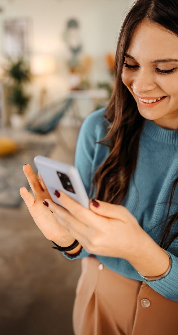 Lächelnde Frau in einem gemütlichen Wohnzimmer, vertieft in ein Smartphone. Ausdruck von Freude und digitaler Kommunikation in entspannter Umgebung.