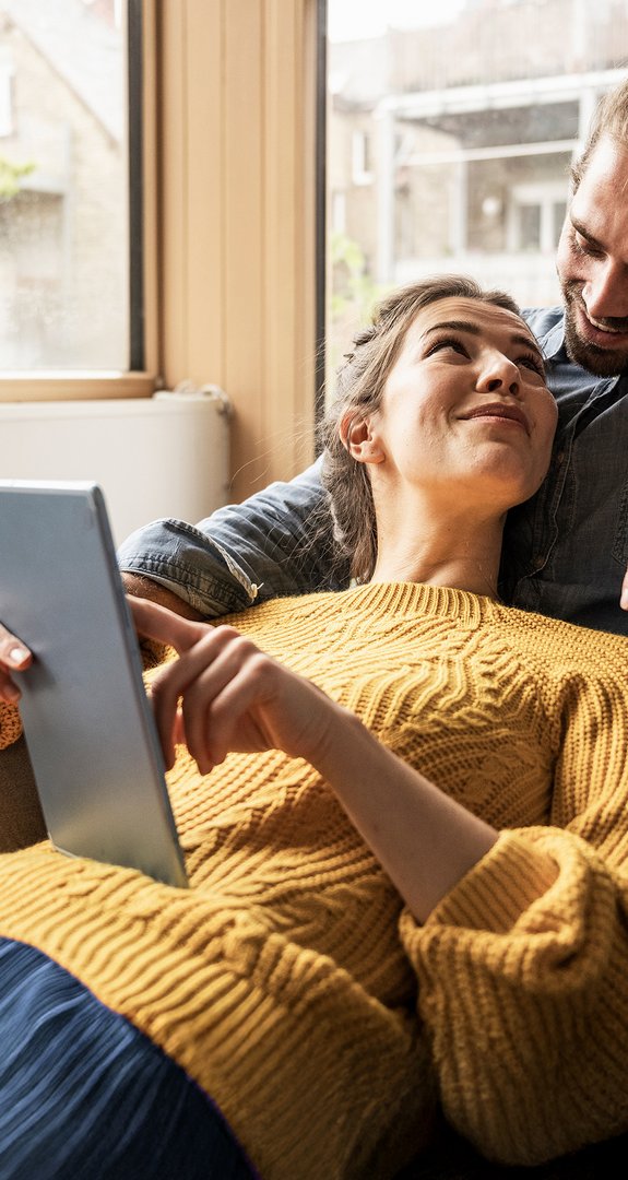 Paar entspannt auf dem Sofa, schaut sich lächelnd an, während eine Person ein Tablet hält.