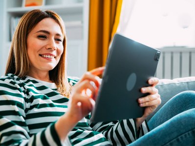 Bild zeigt eine lächelnde Frau mit Tablet auf dem Sofa