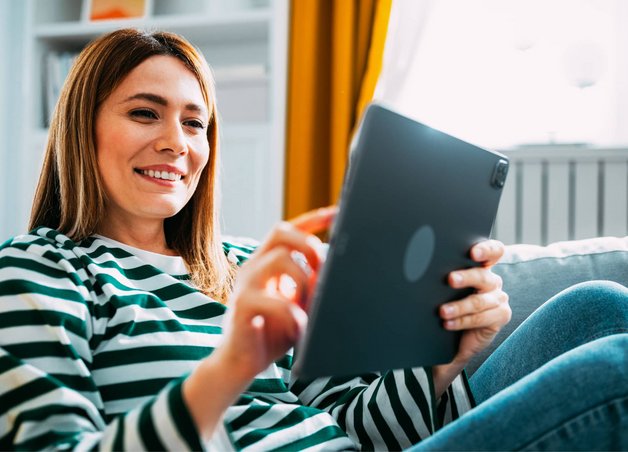Bild zeigt eine lächelnde Frau mit Tablet auf dem Sofa