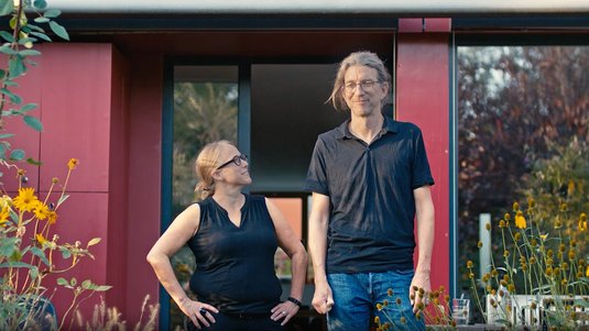 Vorschaubild für ENTEGA-Unternehmensfilm. Zwei Personen stehen vor einem modernen Gebäude mit roter Fassade, umgeben von Pflanzen.
