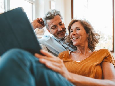 Lächelndes Paar sitzt gemütlich auf dem Sofa, schaut gemeinsam auf ein Tablet. Helle Umgebung, Ausdruck von Entspannung und Gemeinschaft.