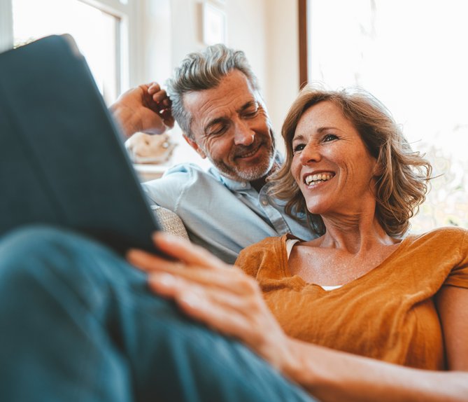 Lächelndes Paar sitzt gemütlich auf dem Sofa, schaut gemeinsam auf ein Tablet. Helle Umgebung, Ausdruck von Entspannung und Gemeinschaft.