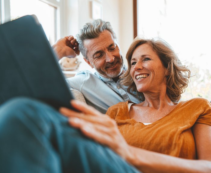 Lächelndes Paar sitzt gemütlich auf dem Sofa, schaut gemeinsam auf ein Tablet. Helle Umgebung, Ausdruck von Entspannung und Gemeinschaft.