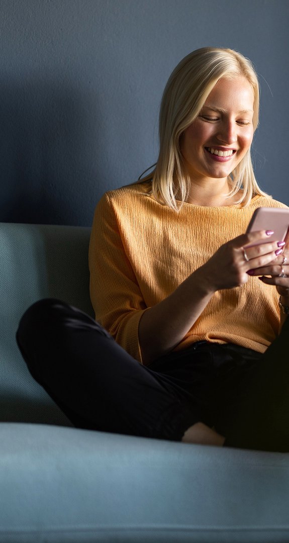Frau sitzt entspannt auf einem blauen Sofa, lächelt beim Blick auf ihr Smartphone. Gemütliche und fröhliche Atmosphäre.