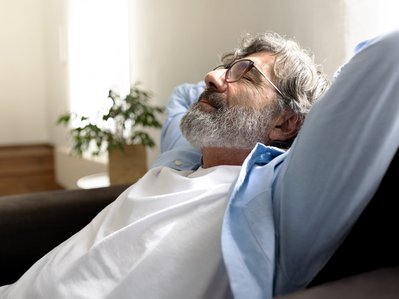 Älterer Mann lehnt sich zurück und entspannt sich auf einem Sofa. Sonnenlicht strahlt durch ein Fenster, im Hintergrund steht eine Pflanze.