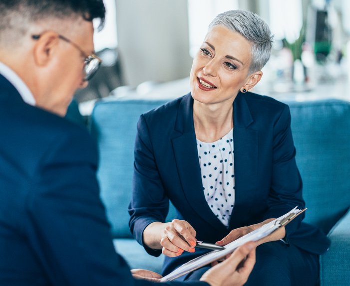 Eine Frau in einem dunkelblauen Blazer spricht aufmerksam mit einem Mann, beide in formeller Kleidung, in einer Büroumgebung.