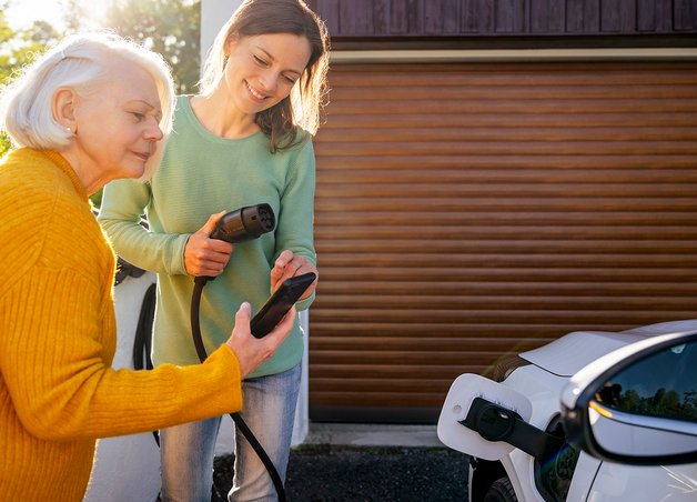 Zwei Frauen laden ein E-Auto an einer Wallbox, informieren sich per Handy über Wallbox Förderung vor einer Garage.