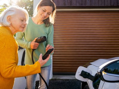 Zwei Frauen laden ein E-Auto an einer Wallbox, informieren sich per Handy über Wallbox Förderung vor einer Garage.