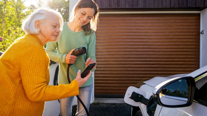 Zwei Frauen laden ein E-Auto an einer Wallbox, informieren sich per Handy über Wallbox Förderung vor einer Garage.