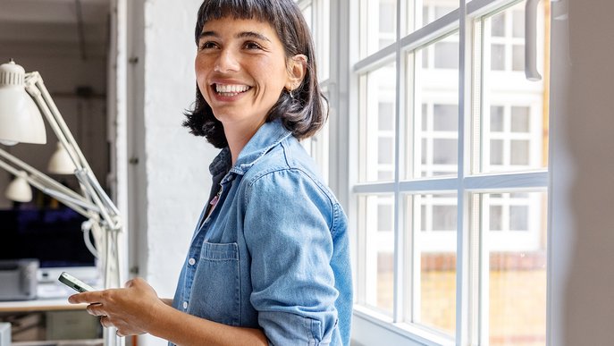 Lächelnde Frau hält Smartphone am Fenster, symbolisiert Zufriedene Kundin mit Smartphone, repräsentiert die effiziente und zugängliche Beratung von ENTEGA.