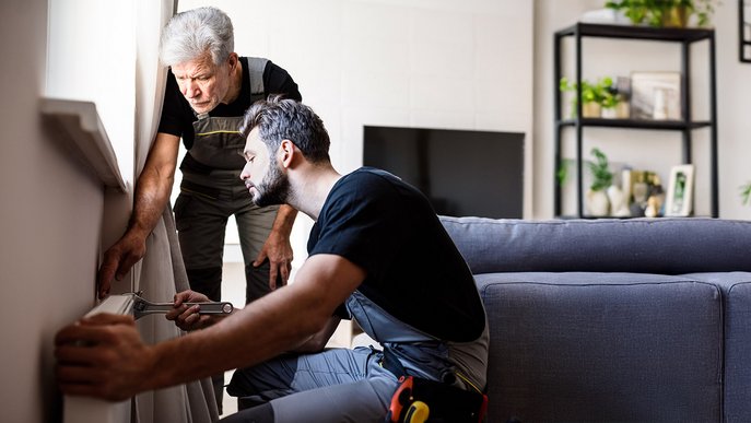 Zwei Handwerker arbeiten konzentriert an einem Heizkörper in einem Wohnzimmer. Der Jüngere kniet mit Werkzeug, ältere Mann beobachtet. Teamarbeit.