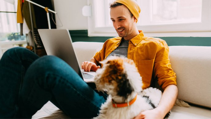 Bild zeigt einen Mann auf dem Sofa, mit Notebook auf dem Schoß und einem Hund daneben