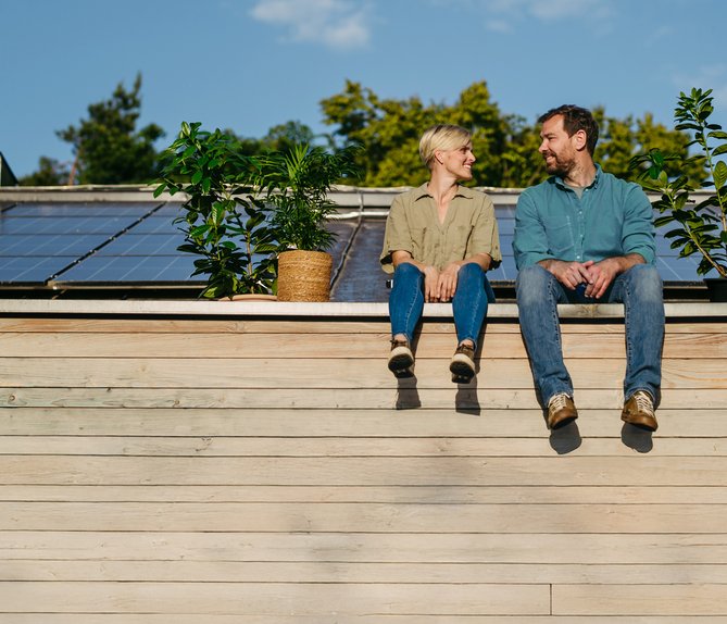Paar sitzt lächelnd vor Solarmodulen auf dem Dach, umgeben von Pflanzen. Darstellung von Unabhängigkeit und Selbstversorgung mit Solarenergie.