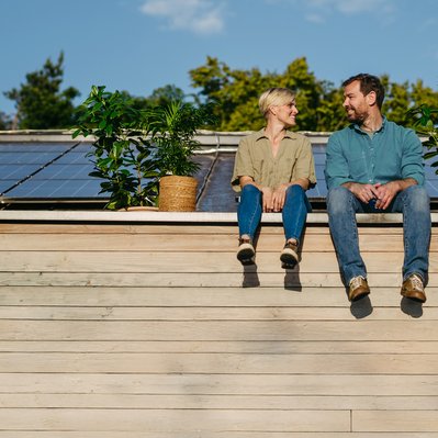 Paar sitzt lächelnd vor Solarmodulen auf dem Dach, umgeben von Pflanzen. Darstellung von Unabhängigkeit und Selbstversorgung mit Solarenergie.