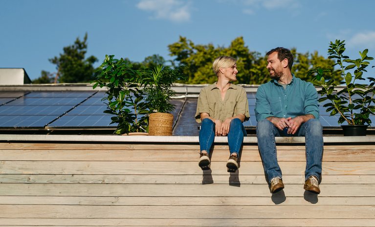 Paar sitzt lächelnd vor Solarmodulen auf dem Dach, umgeben von Pflanzen. Darstellung von Unabhängigkeit und Selbstversorgung mit Solarenergie.