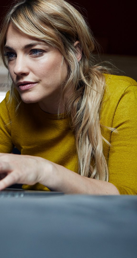 Frau konzentriert sich auf Laptop, trägt gelben Pullover. Zeigt vertieftes Arbeiten oder Surfen in einer komfortablen Umgebung.
