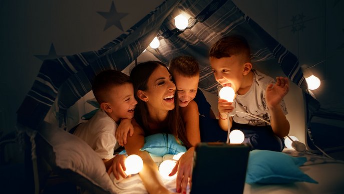 Mutter mit drei Kindern unter einer Decke im Zelt, alle halten leuchtende Glühbirnen, zeigt Freude und gemeinsame Zeit.