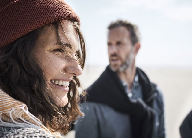 Zwei Menschen stehen im Gespräch am windigen Strand. Beide tragen Mütze und Schal. Sie tauschen sich über die Energiewende in Deutschland aus.
