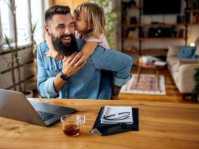 Lachender Mann arbeitet am Laptop, kleines Kind umarmt ihn von hinten. Symbolisiert hilfreiche FAQ-Informationen von ENTEGA zu Glasfaser.