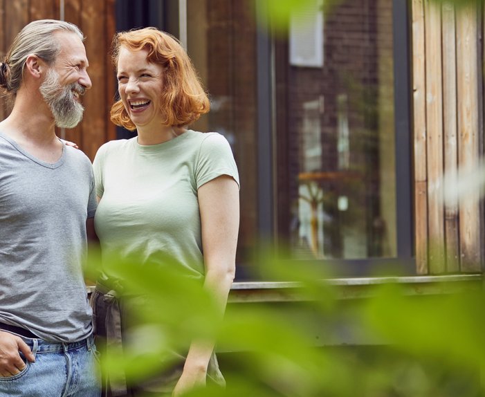 Lächelndes Paar steht vor modernem Holzgebäude, umgeben von Natur. Symbolisiert Energieeinsparung und Klimaschutz durch Gebäudetechnik.