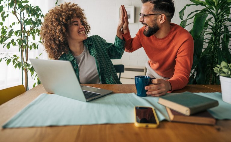 Zwei fröhliche Personen geben sich ein High-Five am Tisch, umgeben von einem Laptop, Büchern und Pflanzen.