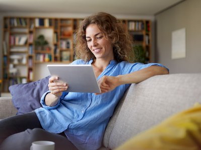 Frau sitzt lächelnd auf dem Sofa im Wohnzimmer und liest auf einem Tablet. Symbolisiert Informationen von ENTEGA zur Zuhause Flat.