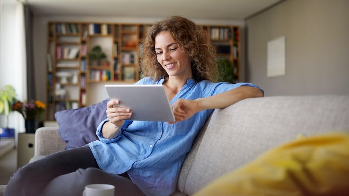 Frau sitzt lächelnd auf dem Sofa im Wohnzimmer und liest auf einem Tablet. Symbolisiert Informationen von ENTEGA zur Zuhause Flat.