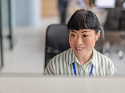 "Eine Frau sitzt nett lächelnd in einem offenen, hellen Büro, schafft eine einladende und kollegiale Atmosphäre. "