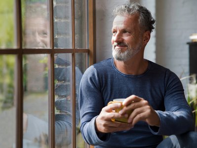 Ein Mann sitzt lächelnd am Fenster und trinkt Kaffee, während er nach draußen schaut. Sein Spiegelbild ist im Glas sichtbar.