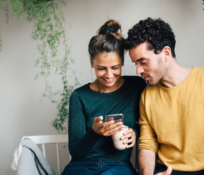 Paar sitzt lächelnd zusammen, schaut auf ein Smartphone. Frau hält eine Tasse. Umgeben von hängenden Pflanzen. Ausdruck von Nähe und digitaler Verbundenheit.