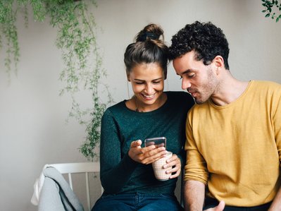 Paar sitzt lächelnd zusammen, schaut auf ein Smartphone. Frau hält eine Tasse. Umgeben von hängenden Pflanzen. Ausdruck von Nähe und digitaler Verbundenheit.