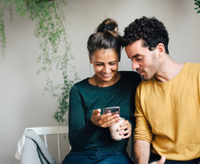 Paar sitzt lächelnd zusammen, schaut auf ein Smartphone. Frau hält eine Tasse. Umgeben von hängenden Pflanzen. Ausdruck von Nähe und digitaler Verbundenheit.