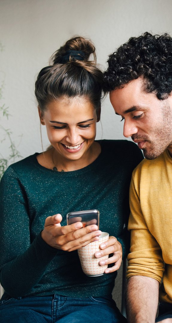 Paar sitzt lächelnd zusammen, schaut auf ein Smartphone. Frau hält eine Tasse. Umgeben von hängenden Pflanzen. Ausdruck von Nähe und digitaler Verbundenheit.