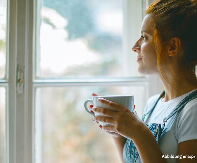 Dank ihrer neuen Kaffeemühle blickt eine Frau mit einer Tasse frisch gebrühtem Kaffee aus dem Fenster und genießt den Moment. 