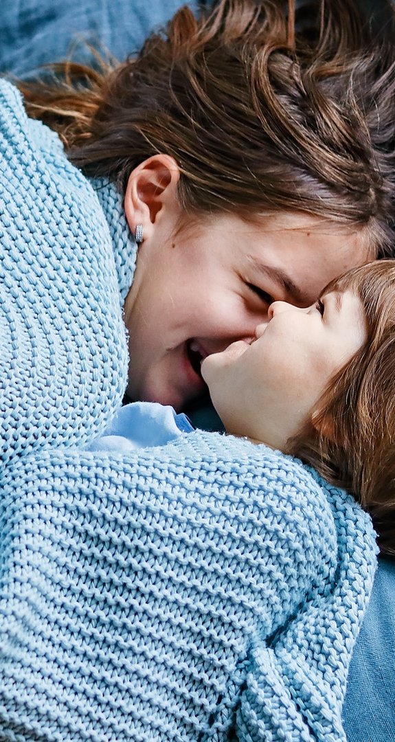 Zwei Kinder kuscheln unter einer blauen Decke auf einem Bett mit blauen Kissen, lächeln sich an und teilen liebevolle, zufriedene Momente.