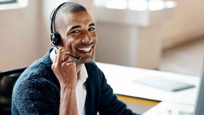 Ein Mann sitzt lächelnd mit einem Headset am Laptop. Symbolisiert fairen Kundenservice.