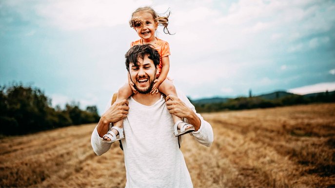 Bild zeigt einen lachenden jungen Vater, der seine fröhliche Tochter auf den Schultern trägt.