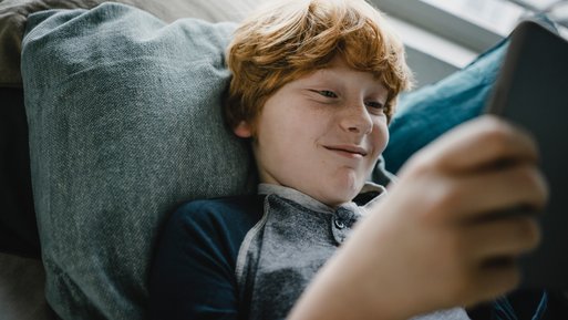 Kind nutzt ein Tablet, gemütlich auf einem Sofa mit Kissen. Ausdruck von digitalem Komfort und nachhaltigem Lebensstil der ENTEGA Zuhause Flat.