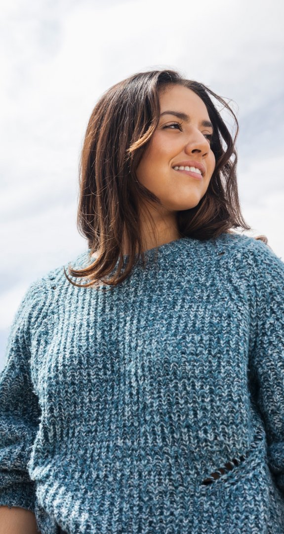 Frau in blauem Pullover lächelt in die Ferne vor einem bewölkten Himmel. Der Ausdruck ist positiv und nachdenklich, vermittelt Freiheit und Leichtigkeit.