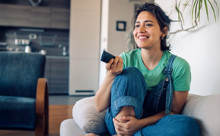 "Lächelnde Person mit Fernbedienung, sitzt zufrieden auf Sofa, symbolisiert entspanntes Fernsehen über ENTEGA TV. "