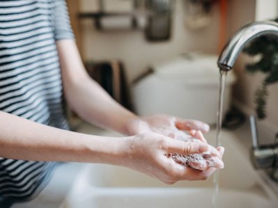 Person wäscht sich Hände am Wasserhahn, Schaum ist sichtbar. Haushaltsszene betont Stromverbrauch vom Durchlauferhitzer für warmes Wasser.