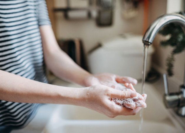 Person wäscht sich Hände am Wasserhahn, Schaum ist sichtbar. Haushaltsszene betont Stromverbrauch vom Durchlauferhitzer für warmes Wasser.