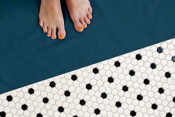 Bild zeigt  auf der einen Hälfte Füße auf einem glatten Boden. Auf der anderen Hälfte grenzt eine Mosaik-Fliesenboden an. Es geht um den Stromverbrauch der Fußbodenheizung.
