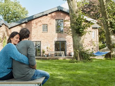 Wärmepumpe Altbau: Außeneinheit neben saniertem Backsteinhaus, Paar genießt Garten, moderne Heiztechnik integriert.