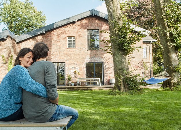 Wärmepumpe Altbau: Außeneinheit neben saniertem Backsteinhaus, Paar genießt Garten, moderne Heiztechnik integriert.